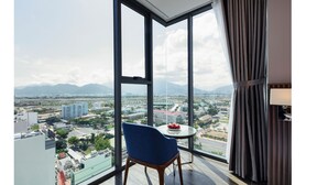 A room with a large window offering a cityscape view, a blue chair, and a small table with fruit. - Amber Hotel (Nha Trang)