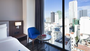 A hotel room with a bed, a blue chair, a small table with fruit, a nightstand with a lamp, and a large window offering a city view. - Amber Hotel (Nha Trang)