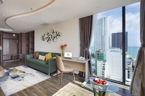 A modern hotel room with a green sofa, a desk with a laptop, a glass table with fruit, and a view of the city and ocean. - Amber Hotel (Nha Trang)