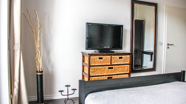A bedroom with a bed, a television on a wooden dresser, a mirror, and a vase with branches. - EXCLUSIVE 2 room apartment with balcony in top location! (Neustadt)