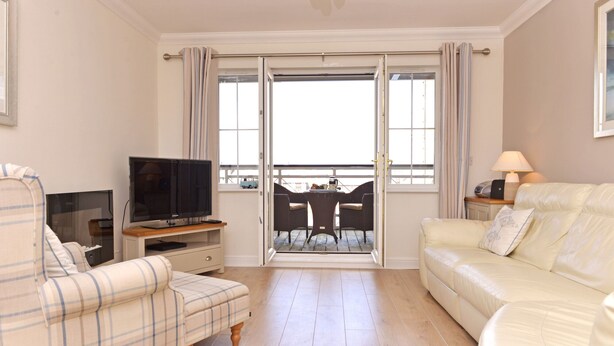 A living room with a sofa, a chair, a TV, a table with chairs, and a balcony with curtains. - Pacific Heights (Eastbourne)