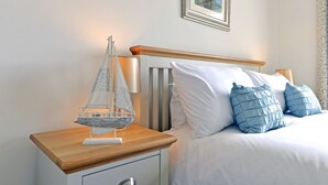 A neatly made bed with white and blue bedding, a wooden nightstand with a model sailboat, and a lamp. - Pacific Heights (Eastbourne)