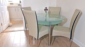 A round glass table with four beige chairs, a vase with purple flowers, and a kitchen in the background. - Pacific Heights (Eastbourne)