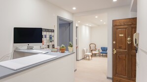 A reception desk with a computer monitor, keyboard, and notepad. There are potted plants and a sign on the wall. A wooden door and a blue chair are visible in the background. - Santo Spirito Venti (Florence)