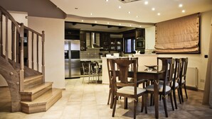 A modern kitchen with dark wooden cabinets, a stainless steel range hood, and a dining area with a wooden table and chairs. - Villa Theano - 4 bedroom villa close to the beach (Agios Nikolaos)