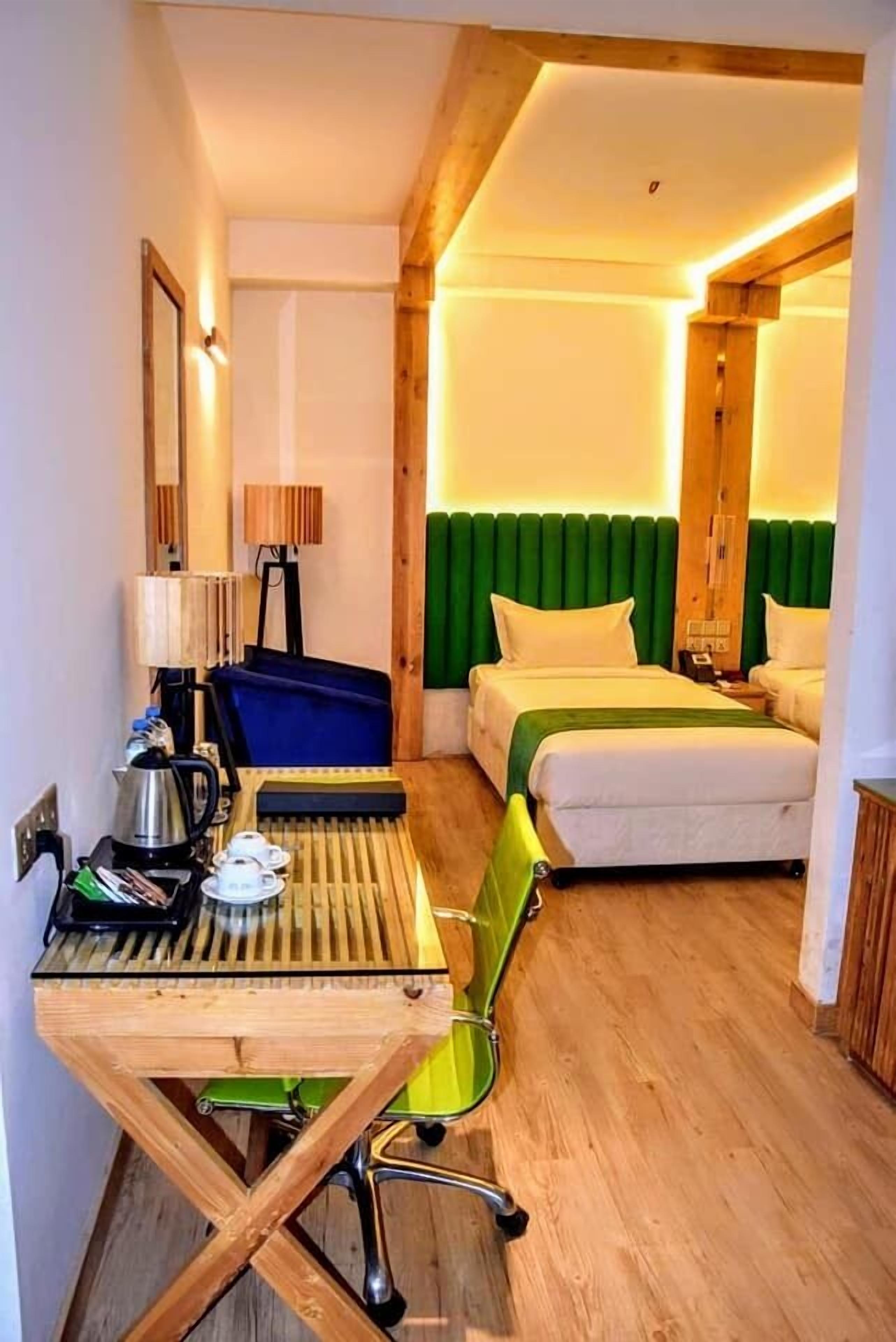 A hotel room with a wooden desk, a bed with green headboard, and a small table with a kettle and cups.