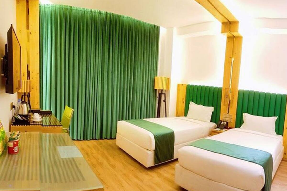 A hotel room with two beds, a wooden table, a green curtain, and a lamp.