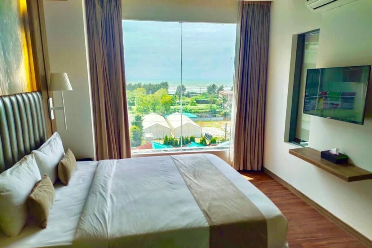 A hotel room with a bed, a TV on the wall, and a window with a view of greenery and buildings.