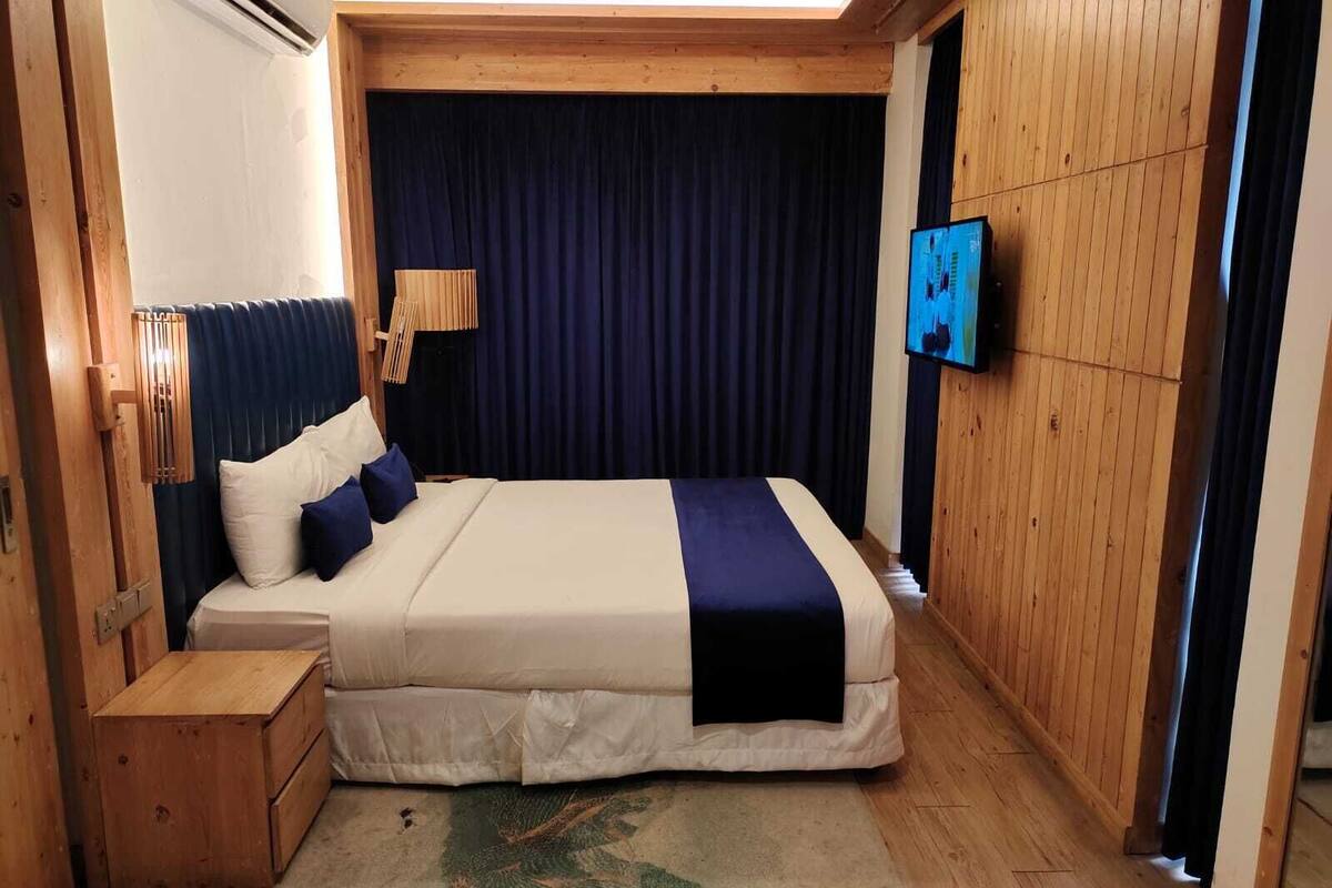 A bedroom with a bed, wooden headboard, a nightstand, a TV mounted on the wall, and a window with curtains.