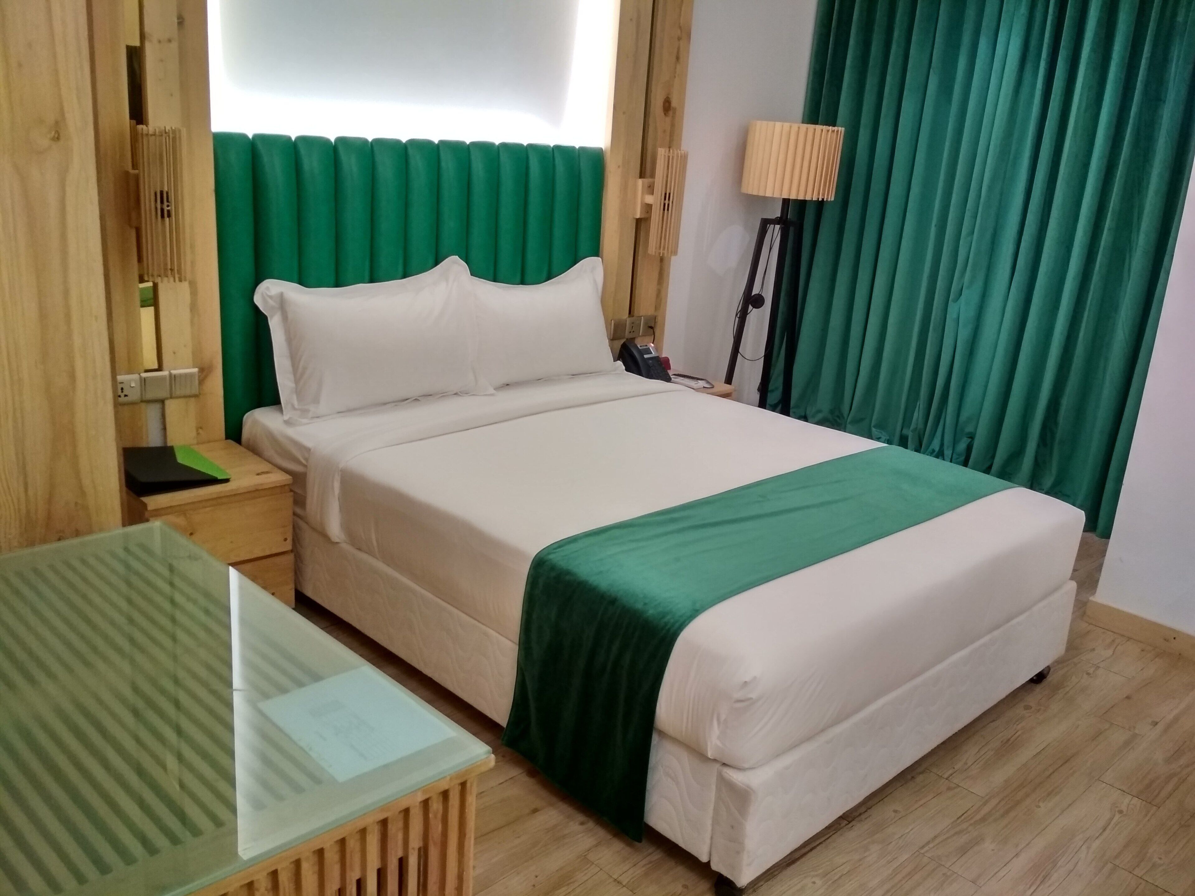 A neatly arranged hotel room with a bed, a bedside table, a lamp, and a green curtain.
