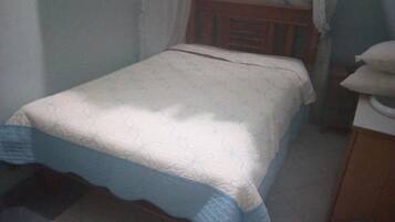 Une petite chambre avec un lit simple, une tête de lit en bois, une couvre-lit blanche et bleue, et une table de chevet avec une lampe.