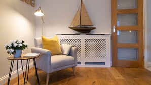 Una habitación con piso de madera, un sillón gris con un cojín amarillo, una mesita con una planta en maceta, una decoración de velero colgado en la pared y una puerta de madera con paneles de vidrio esmerilado.