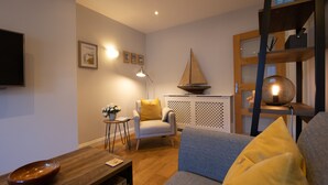 Una sala de estar moderna con una mesa de centro de madera, un sofá gris, una pequeña mesita de noche con una planta, un sillón blanco y una estantería con un modelo de velero.