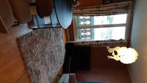 Een kamer met een houten vloer, een kamerbreed tapijt met dessin, een bureau met stoel, een raam met gordijnen en een hanglamp.