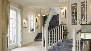 A spacious hallway with a staircase leading to a second floor. The walls are adorned with framed pictures and wall sconces. - Hotel Buchenhof (Worpswede)