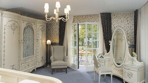 A room with a large bed, a chair, a dresser with a mirror, and a chandelier. There is a window with curtains and a view of the outdoors. - Hotel Buchenhof (Worpswede)