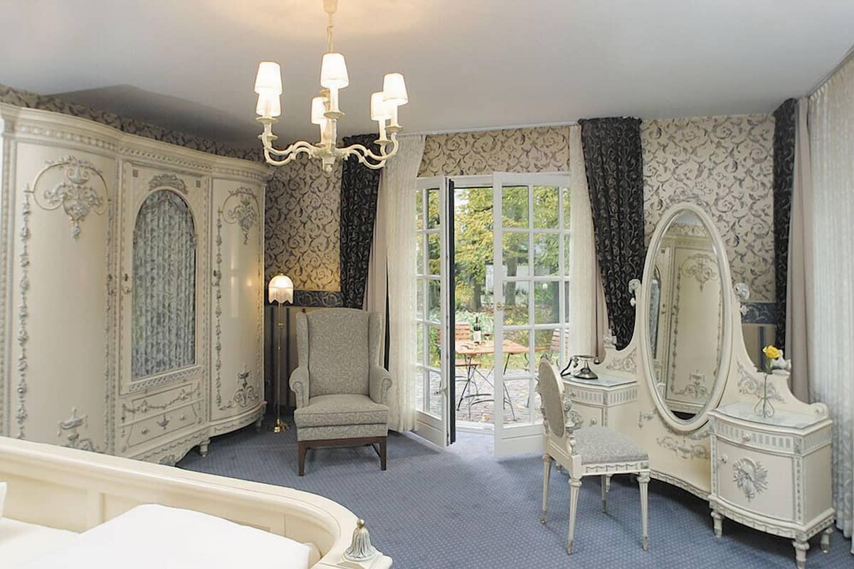 Ein Zimmer mit einem großen Bett, einem Sessel, einer Kommode mit Spiegel und einer Chandelier. Ein Fenster mit Vorhängen bietet Blick ins Freie.