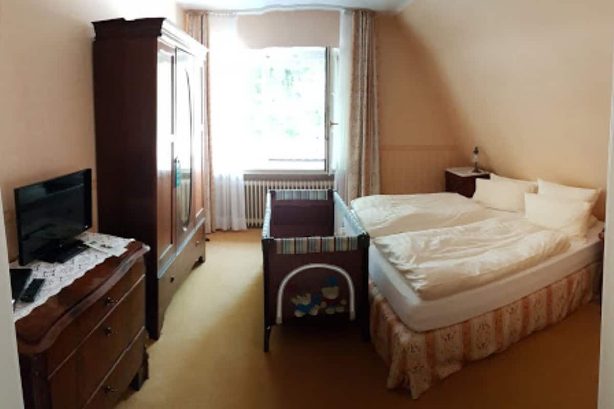 Ein Schlafzimmer mit einem Bett, einer Kommode, einem Fernseher, einem Fenster mit Vorhängen und einer Heizung.