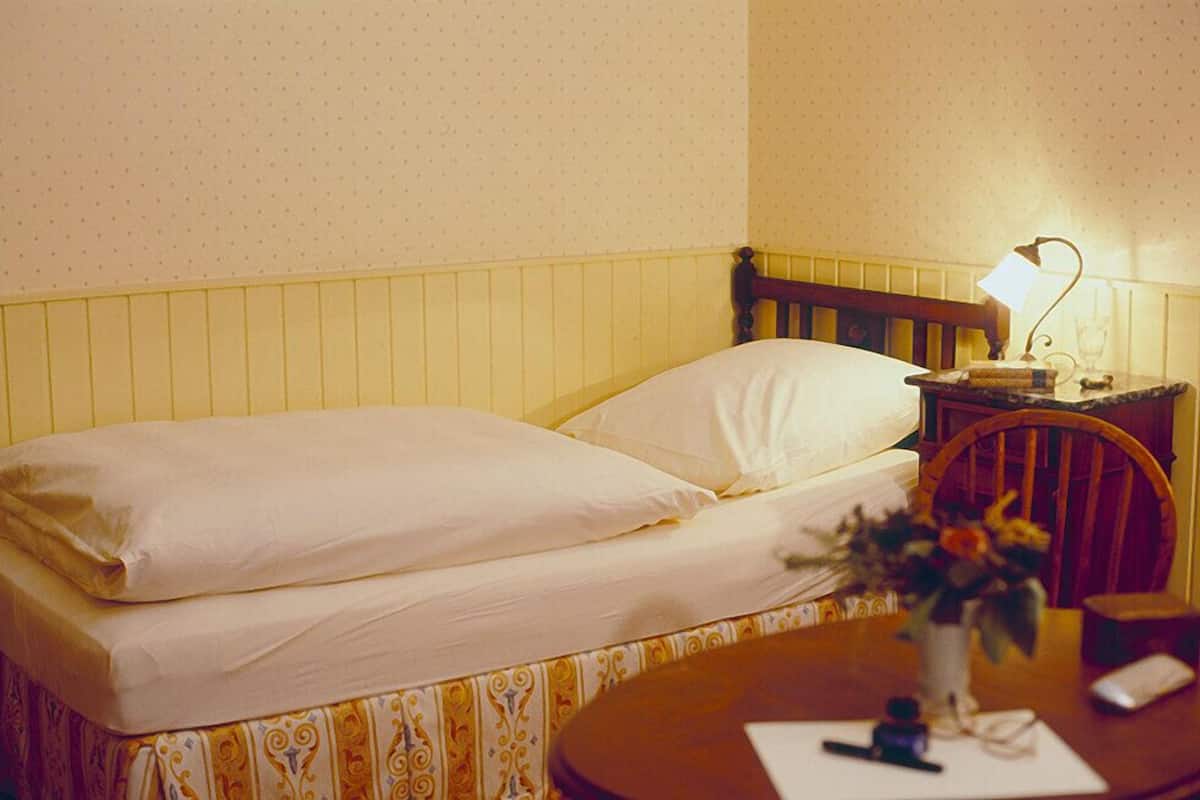 Ein Einzelbett mit hölzernem Kopfteil, ein Nachttisch mit Lampe und ein kleiner Tisch mit Blumenvase.