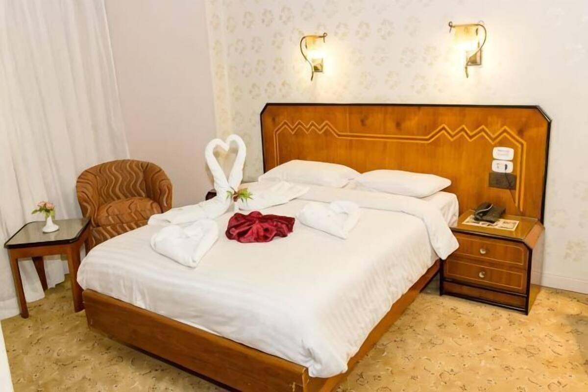 A hotel room with a wooden bed, two wall-mounted lamps, a nightstand, a chair, and a small table with a flower.