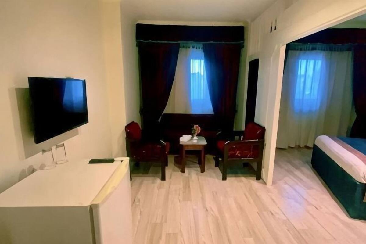 A living room with a flat-screen TV, a small table, two chairs, a sofa, and a bed.
