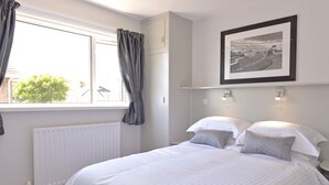 A bedroom with a large bed, a window with curtains, a radiator, a framed picture on the wall, and a shelf with two wall-mounted lights. - Beach Garden (Pevensey)