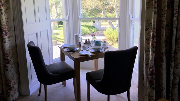 A room with a bay window overlooking a garden, featuring a table set with tea cups and a kettle. - The Burrows Bed and Breakfast (Pembroke)