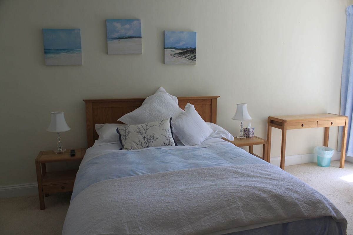 A bedroom with a bed, two bedside tables, a nightstand, a wooden side table, and three paintings on the wall.