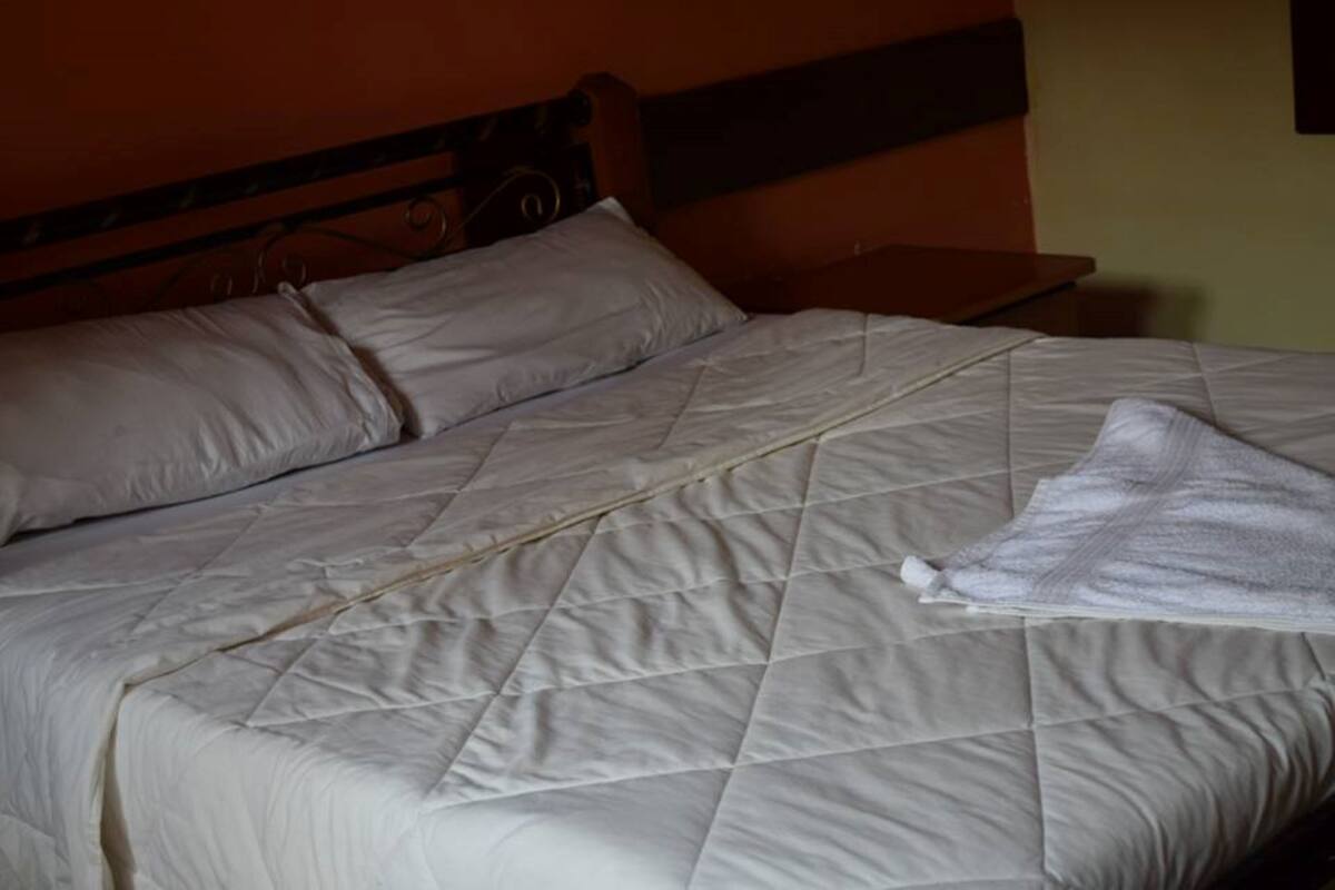 Una cama con edredón blanco acolchado y almohadas, cabecera de madera y mesita de noche con lámpara.