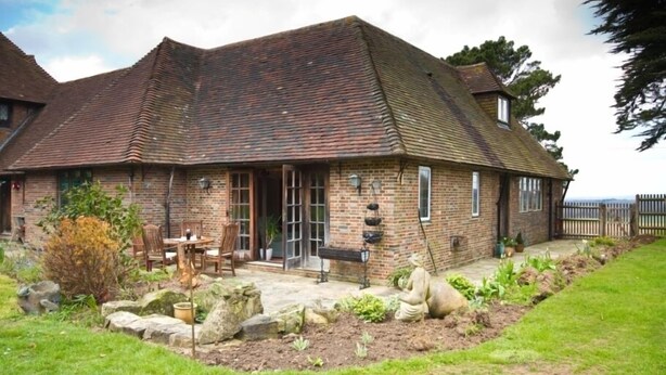 A traditional brick house with a sloped roof, a front porch with chairs, and a garden with rocks and plants. - Cottage at Friston Down (Eastbourne)
