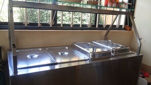 Disposição de buffet em aço inoxidável com três recipientes tampados, uma prateleira de vidro acima e uma janela com grade metálica.