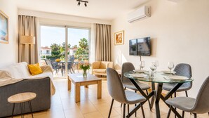 A modern living room with a sofa, a dining table with chairs, a TV mounted on the wall, and a balcony with a view. - Narcissos 'Nissi Beach' Apartment C3 (Ayia Napa)