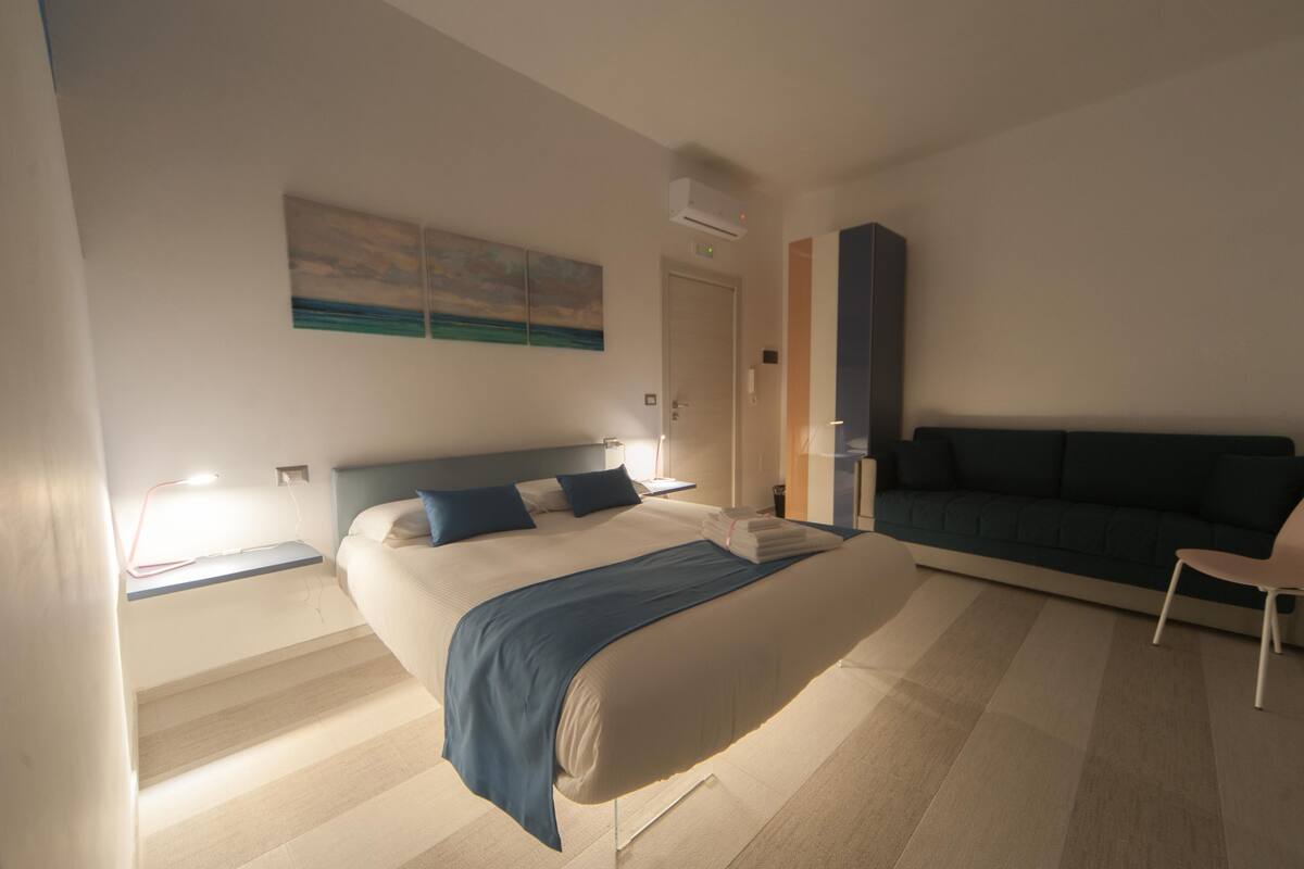 Quarto de hotel moderno com uma cama grande, um sofá, uma escrivaninha com abajur e um quadro na parede.