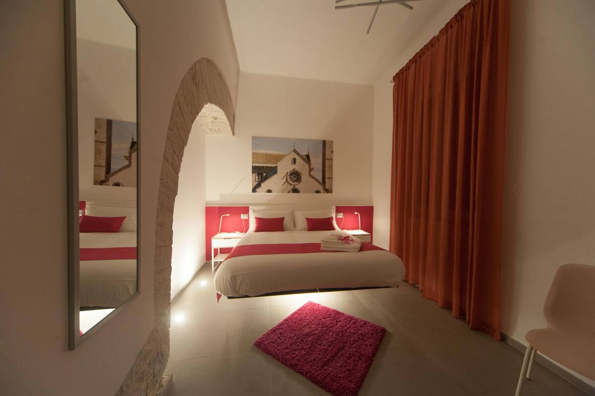 Quarto de hotel moderno com uma cama grande, uma parede de destaque vermelha, um espelho amplo e um quadro de um edifício.