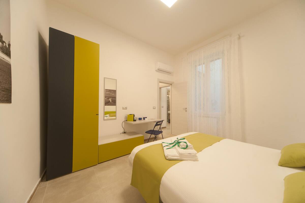 Quarto de hotel com cama, escrivaninha, cadeira e banheiro visível por uma porta aberta.