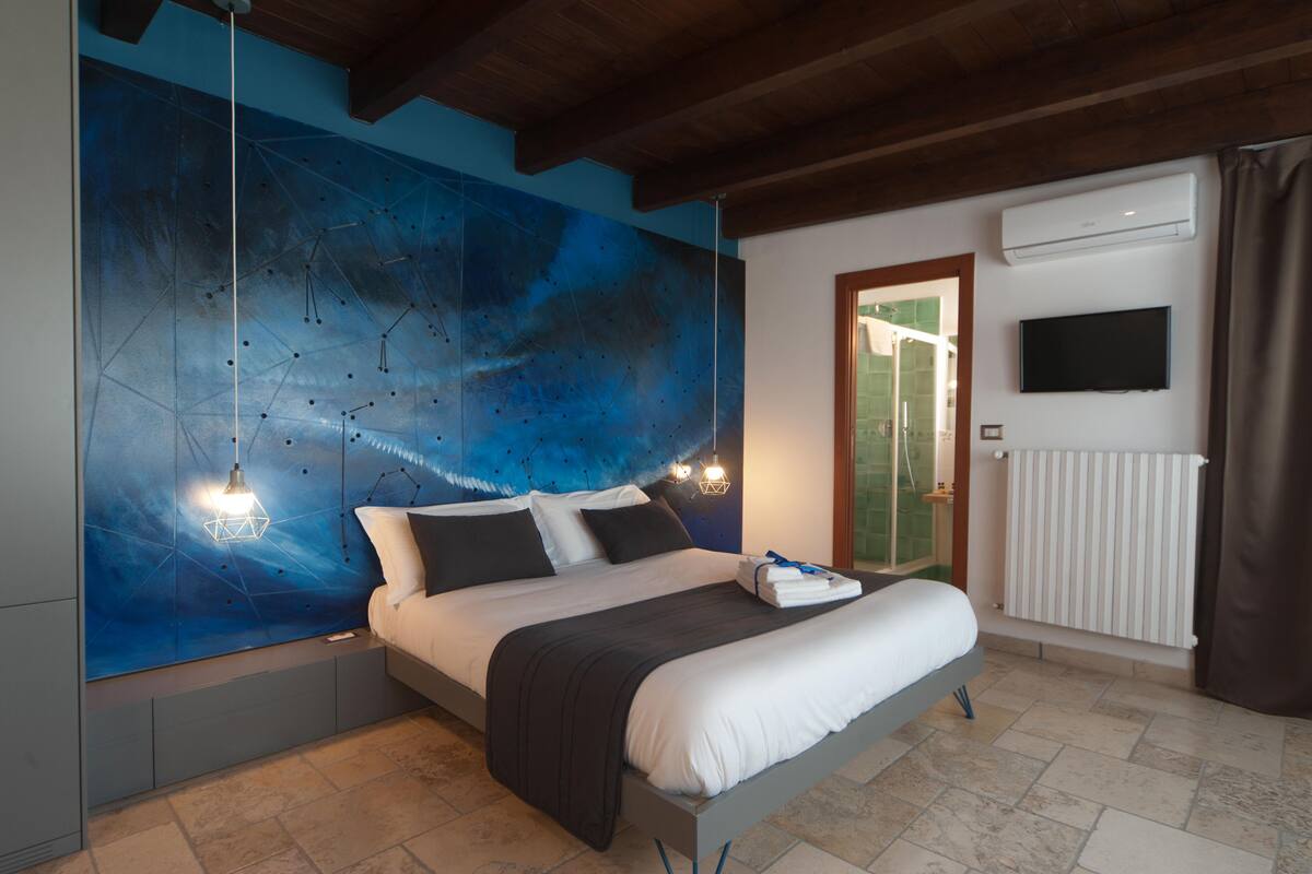 Quarto moderno com uma cama grande, parede azul abstrata, televisão montada e uma janela com cortinas.