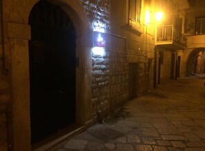 A narrow stone-paved street with a closed arched doorway and a lit sign on the wall. - La Cattedrale dai Tetti (Trani)