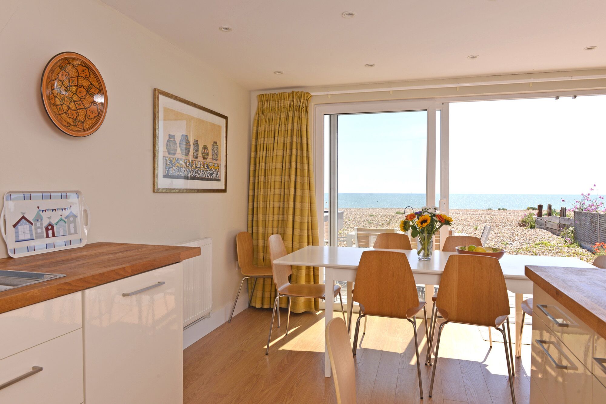 A dining area with a view of the beach, a table with chairs, a vase of flowers, and a plate with a design on the wall.