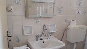 Ein Badezimmer mit einem weißen Waschbecken, einem Spiegel darüber und einer Toilette. Handtücher liegen auf einem Regal und ein Mülleimer steht bereit.