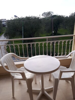 A balcony with a round table and two chairs, a railing, and a view of a road and greenery. - Sophie Studios (Corfu)