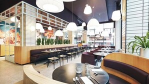 A modern restaurant interior with round tables, cushioned seating, pendant lighting, and a large window with a view. - Kinley Cincinnati Downtown, a Tribute Portfolio Hotel (Cincinnati)