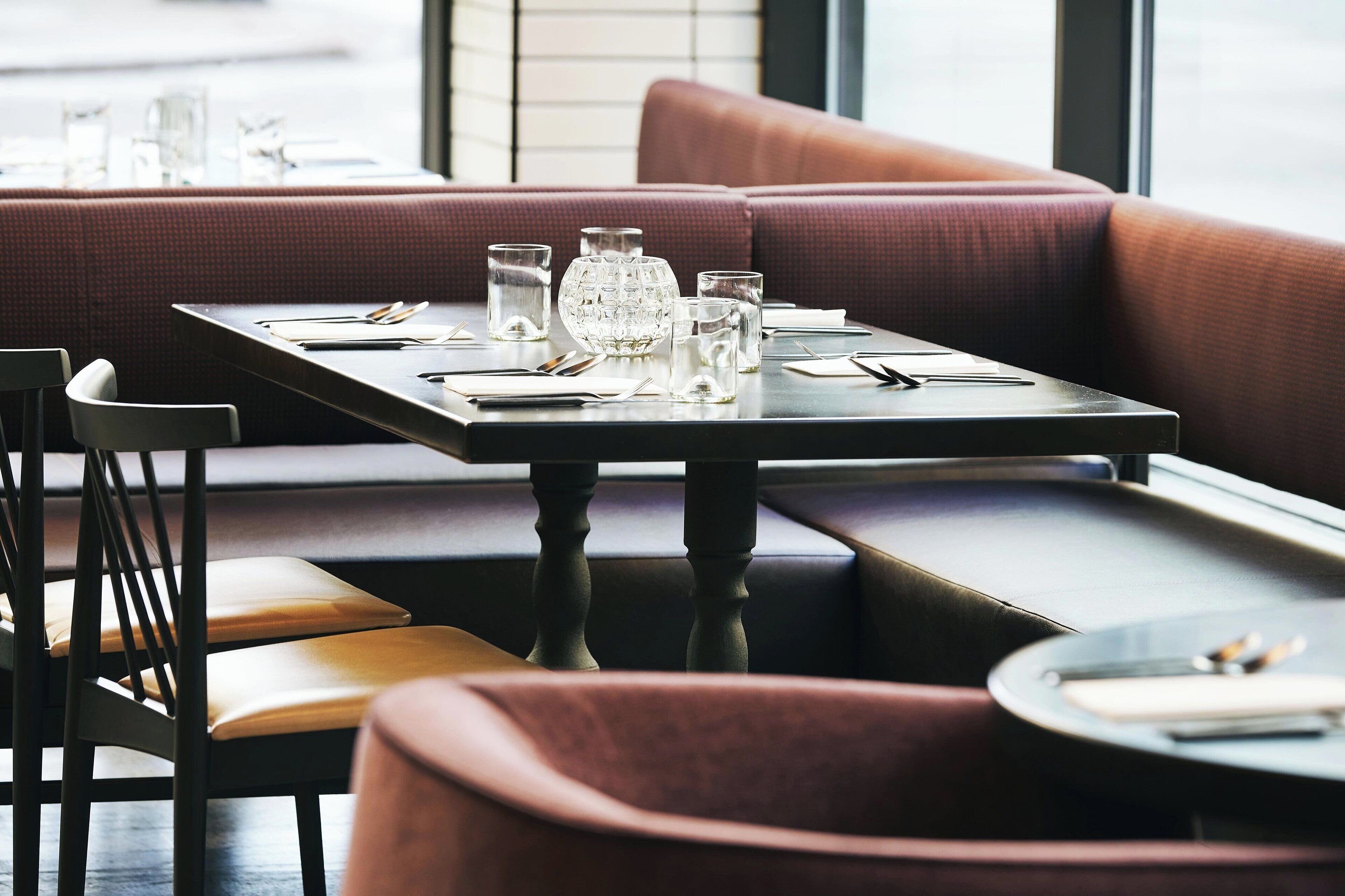 A restaurant with a booth seating area, a table set with cutlery and glasses, and a window view.