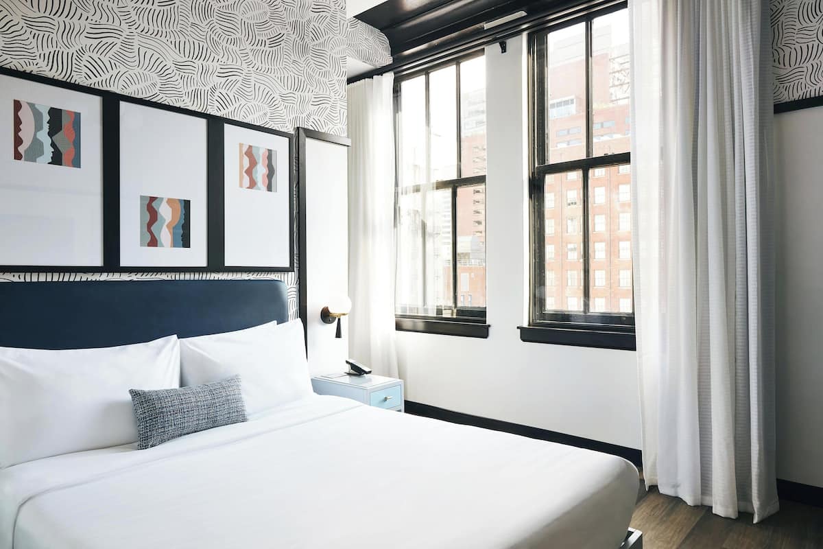 Hotellihuone, jossa on suuri sänky, yöpöytä, ikkuna verhoilla ja taideteos seinällä.