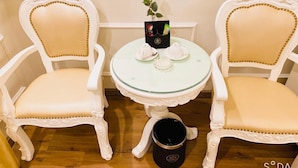 A corner with two white upholstered chairs with gold beading, a small round table with a glass top, and a red rose on the table.