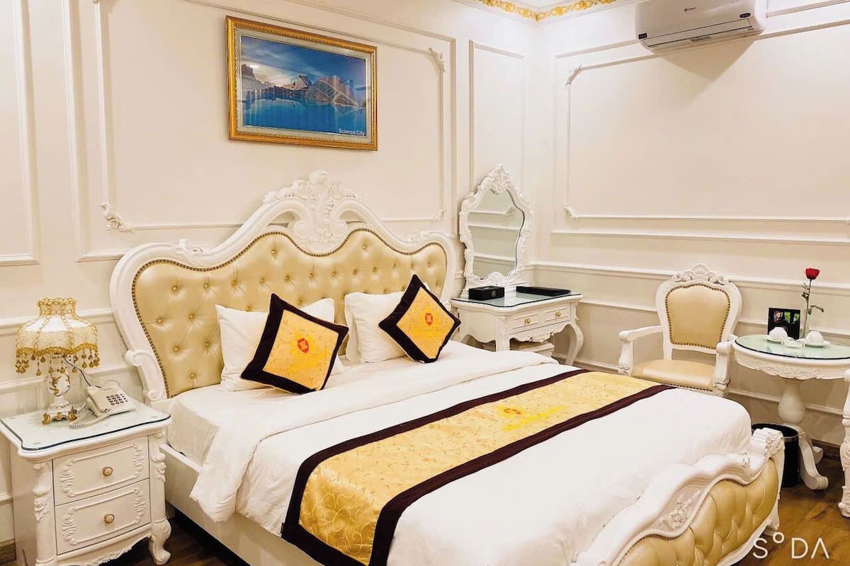 A hotel room with a large bed, a bedside table with a lamp, a chair, a small table with a vase, and an air conditioning unit on the wall.