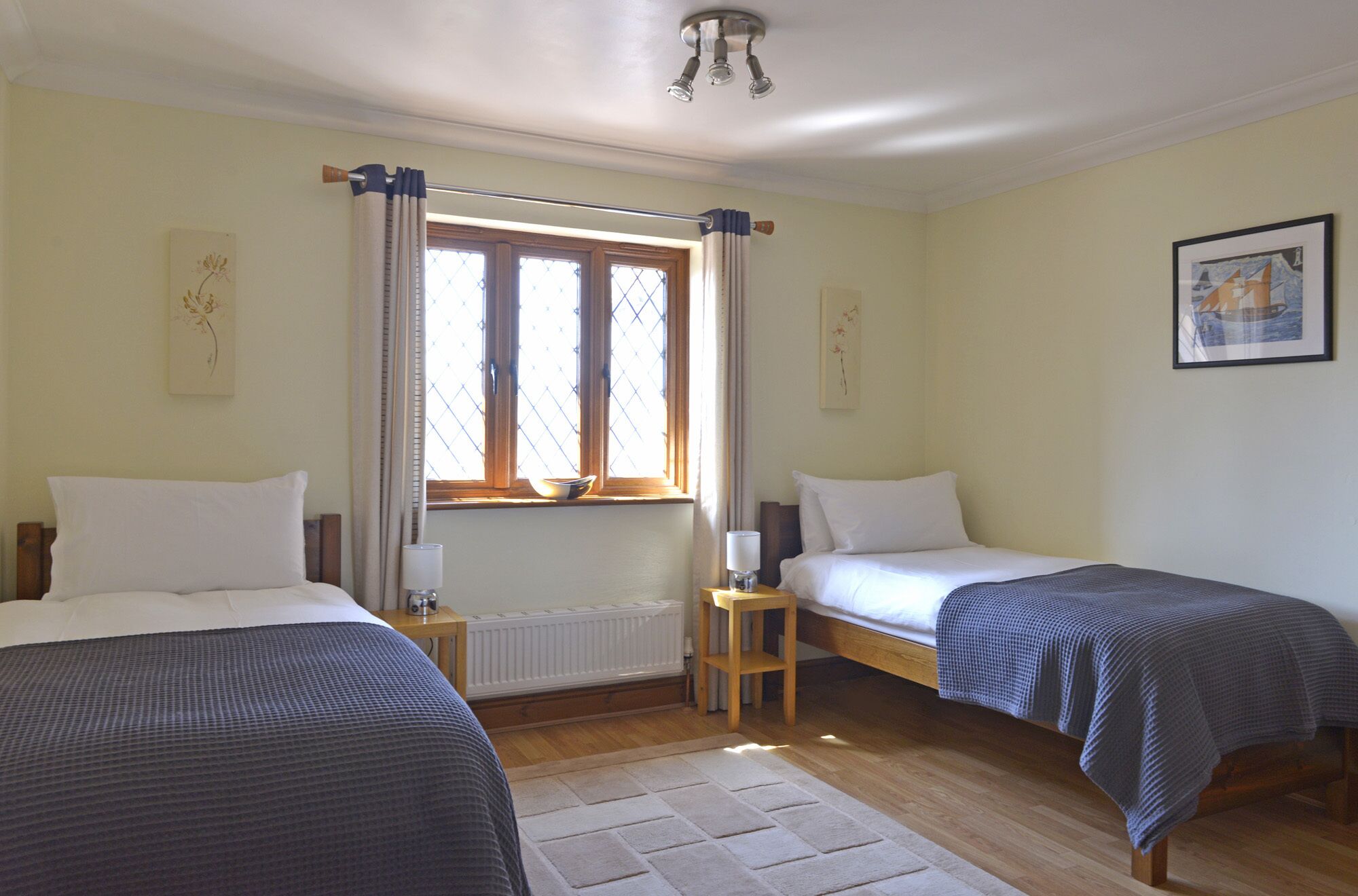 Una habitación con dos camas individuales, una ventana con cortinas, un radiador, una mesita con una lámpara y un cuadro en la pared.