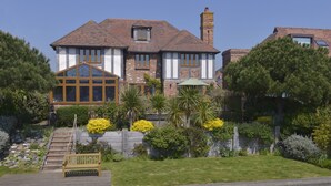 Una casa de estilo Tudor con un amplio ventanal, un invernadero y un jardín cuidado con una banca y diversas plantas.