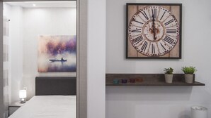 A hotel room with a bed, a painting of a boat on water, a clock, and a shelf with plants. - InCity La Central 203, Deluxe Flat (Thessaloniki)