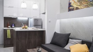 A modern kitchen with white cabinets, a central island, and pendant lighting. There is a dark-colored sofa with cushions and a small table with a glass container. - InCity La Central 203, Deluxe Flat (Thessaloniki)