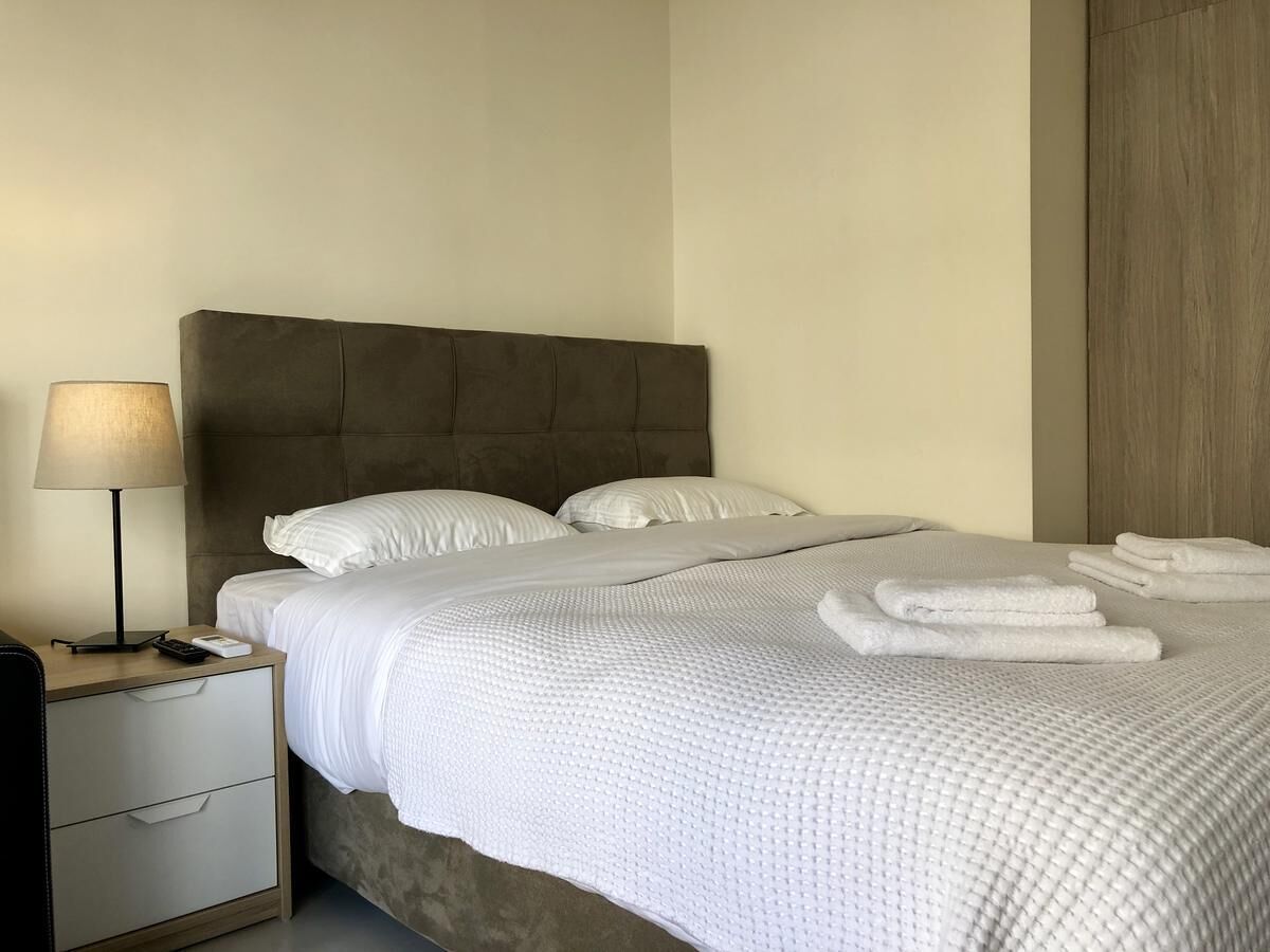 Una cama bien tendida con ropa blanca y toallas dobladas, una lámpara de noche y una mesita de noche de madera con un control remoto.